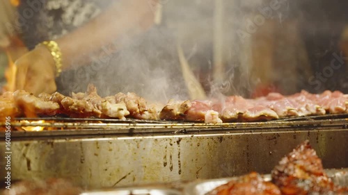 Slow Motion Vendor Turning Pork Skewers Over Flaming Smoky Grill at Night Market, close angle, Street Food Concept