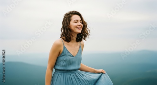 A young woman in a blue dress standing on a mountain top with her eyes closed, enjoying the view.