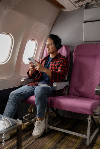 Asian man sitting comfortably on airplane seat near window, holding smartphone and passport, wearing headphones around neck, enjoying in-flight entertainment. Modern travel lifestyle and technology.
