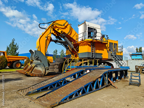 Large yellow excavator with a powerful digging arm, positioned on a construction site, surrounded by machinery and blue sky, showcasing heavy equipment in action