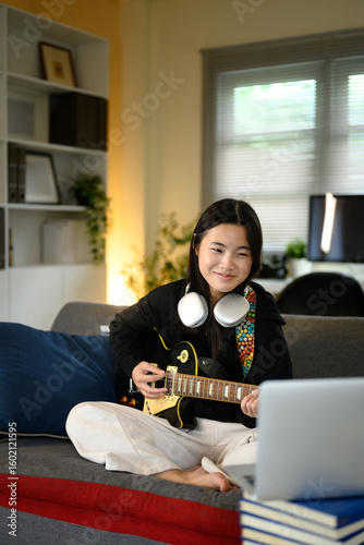 Teenage girl learning to play electric guitar at home and watching an online lesson on laptop