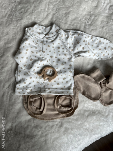 A turtleneck, shorts, socks and a wooden elephant rodent lie on the light blanket of the bed in the room. A set of clothes for discharge for a newborn baby. Top view