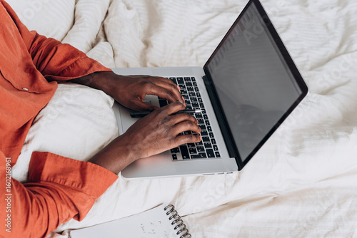 Freelancer African American is working from home, typing on a laptop while sitting in bed, with a notebook and pen nearby, enjoying the flexibility and comfort of remote work