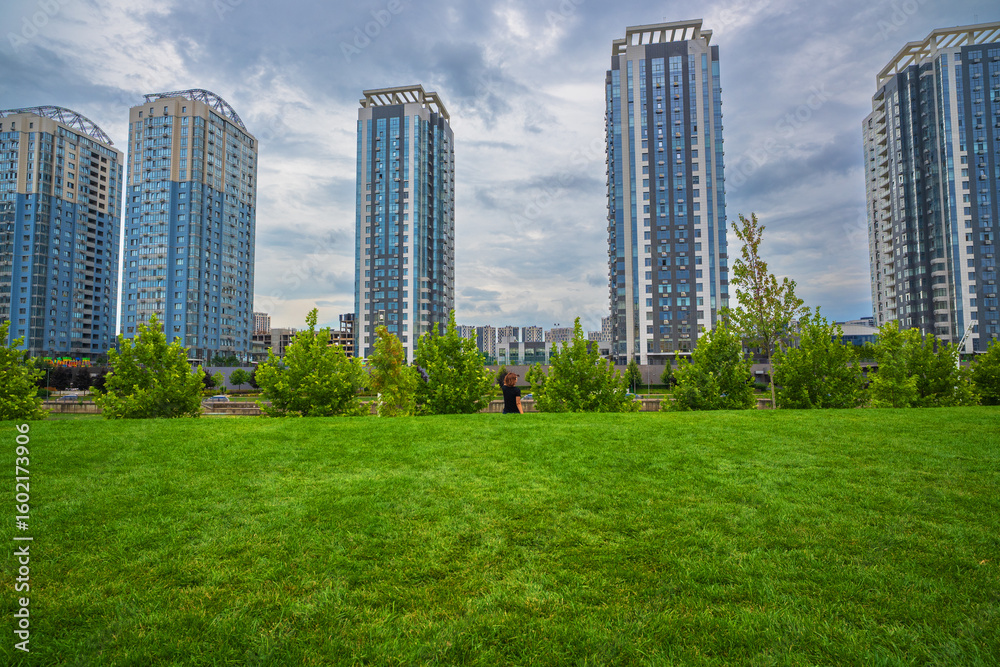 Obraz premium Modern high-rise residential buildings stand tall behind a vibrant green lawn and lush trees under a cloudy sky, showcasing contemporary urban living with refreshing natural elements.
