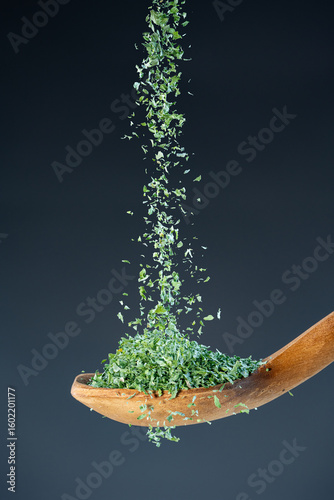 Close-up of crushed parsley or herb flakes fall from above onto a wooden spoon, ideal for culinary and food photography concepts.