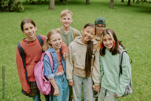 Group of diverse children standing together in park smiling at camera, multiethnic boys and girls with backpacks posing outdoors during school outing or educational activity