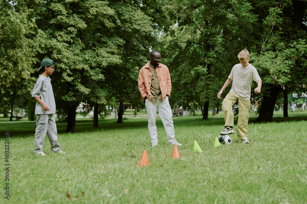 Obraz premium Diverse Boys playing soccer with cones on grass in park with young Black male teacher, engaging in outdoor activity together
