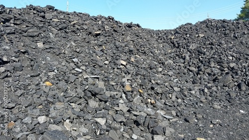 A large pile of dark coal fragments and rocks under a clear blue sky, showcasing a rough, uneven surface typical of a mining or industrial site
