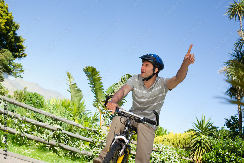 Fototapeta premium Mężczyzna w kasku rowerowym jadący rowerem po utwardzonej ścieżce w parku z ogrodzeniem i tropikalnymi krzewami