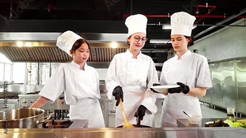 Chef instructor and students in uniform plate freshly cooked pasta in a commercial kitchen. The scene shows teamwork and hands-on learning, ideal for culinary school or food preparation concepts.