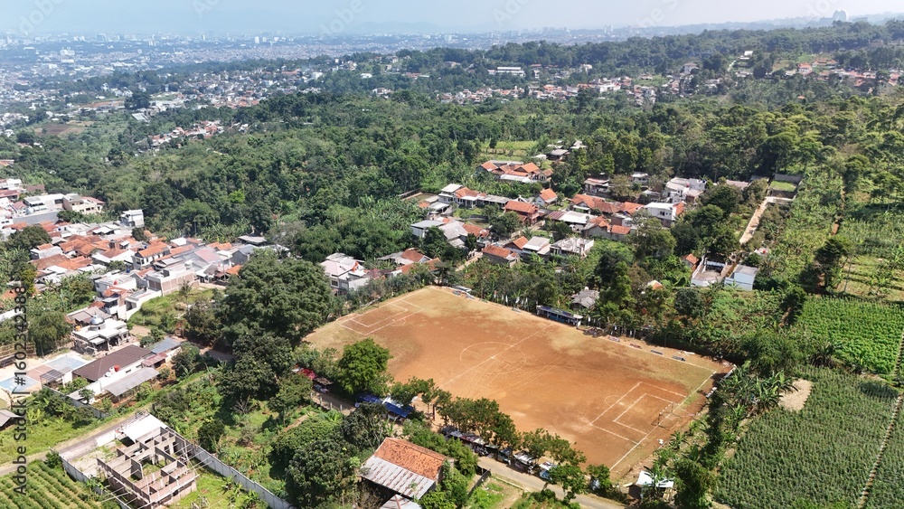 Fototapeta premium Bandung, July 23, 2025: Aerial shot of Lebak Gede football field in Cimenyan, Bandung. Surrounded by green forest, farmland, and traditional village houses near the city edge.
