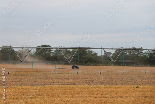 Agricultural field with central pivot irrigation system and tractor in background. Dry grass and dust in air during automated watering on farmland, modern farming technology concept.

