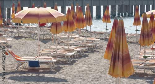 Ombrelloni da spiaggia arancioni e gialli con lettini vuoti sulla costa del Mediterraneo con pontile sullo sfondo.