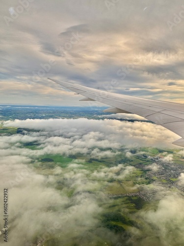 An aerial view from an airplane window showing a wing stretching over a dramatic layer of clouds, with green fields and scattered towns visible below and a softly glowing sky above.