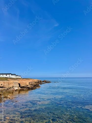 A peaceful coastal scene featuring clear blue water gently meeting rugged rock formations, with a row of white cottages perched near the edge under a bright blue sky.
