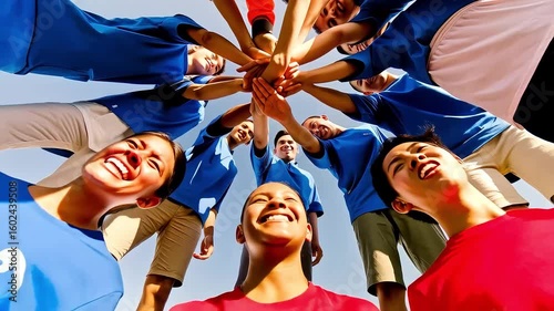 A group of young people in athletic wear huddle, stacking hands and smiling up, symbolizing unity and teamwork against a bright sky