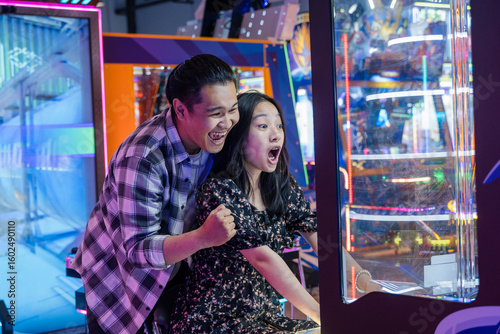 Excited young Asian couple playing an arcade game together in a neon-lit game center, both showing expressive reactions of joy and surprise while looking at the screen in a fun indoor atmosphere
