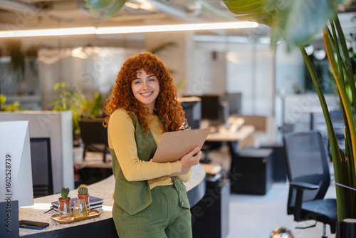 Фотография Young businesswoman holding clipboard smiling in modern green office