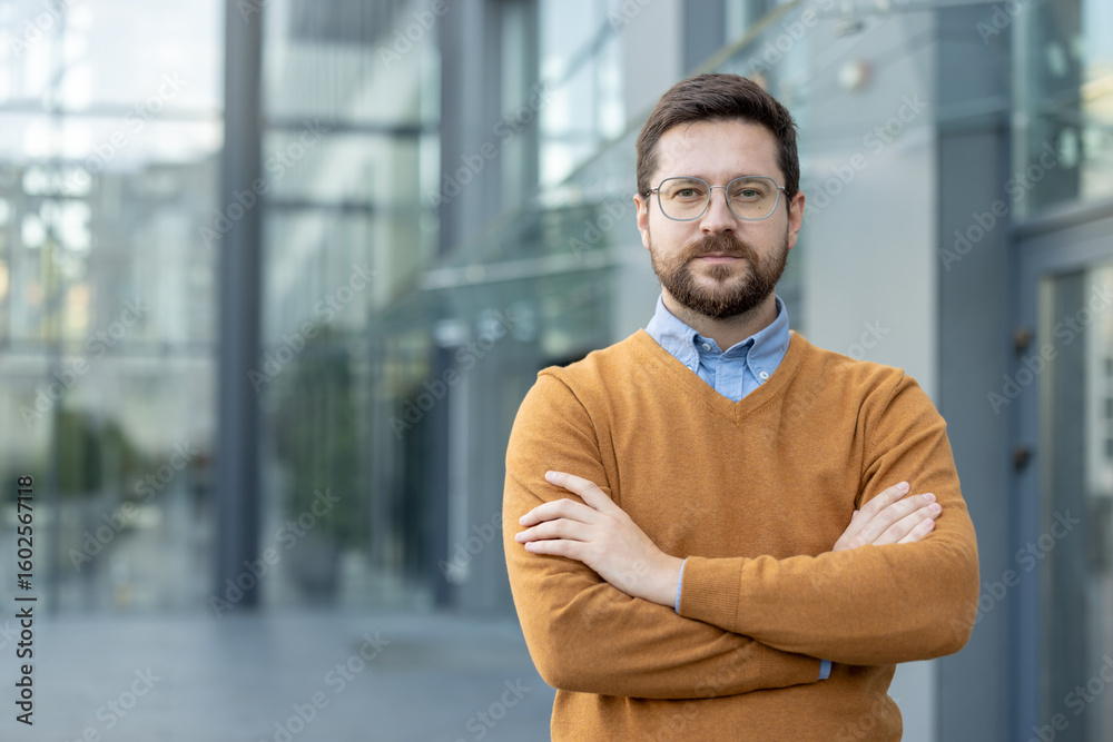 Obraz premium Close-up portrait of a serious young confident businessman standing on the street near his office, arms crossed on his chest and looking at the camera.