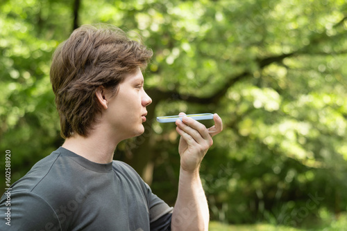 Young caucasian blond man recording voice message on cellphone. Outdoors. Close up