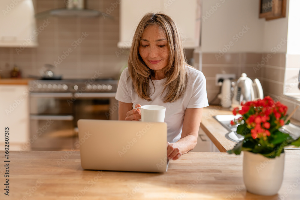 Woman enjoying coffee while working on laptop in a cozy kitchen setting during a sunny morning Woman enjoying coffee while working on laptop in a cozy kitchen setting during a sunny morning