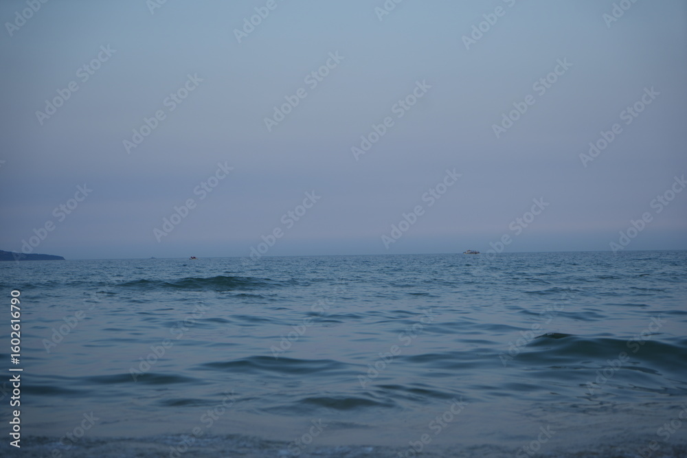 Obraz premium Calm sea at dawn with soft pastel blue and pink tones. Gentle waves approaching the shore under a peaceful, minimalistic horizon.
