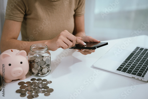 An Asian woman is writing in a notebook and stacking coins and a piggy bank, which symbolizes budgeting, financial planning, saving money and smart money management.