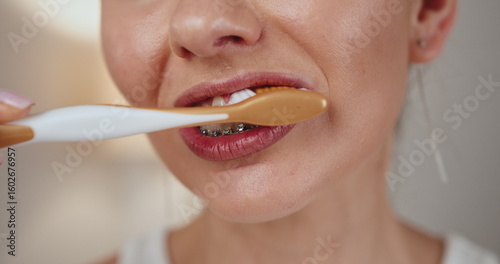 Young Caucasian woman brushing teeth with toothbrush, focusing on dental care and oral hygiene. Braces are visible, emphasizing dental health and orthodontics. Image captures routine morning dental