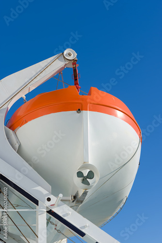 A bright orange and white lifeboat, secured by a davit system, is seen from a low angle against a soft, evening sky, highlighting its readiness on a vessel.