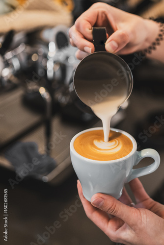 Barista pouring steamed milk into espresso, creating latte art in a heart shape. Close-up of hands and cup on a countertop. Morning coffee preparation in a café setting.