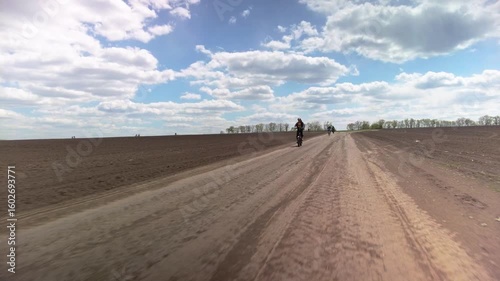 Healthy athletic people riding bikes through soil fields. Group of cyclists approaching on wide dirt road, open fields and blue sky with clouds around.