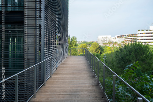 Contemporary footbridge blending wooden textures and industrial architecture, flanked by metal facades and green surroundings in an urban renewal area