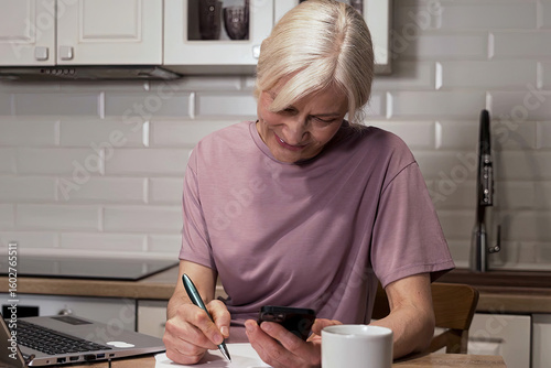 Focused Senior Woman Engaged in Multi-tasking with Phone and Notepad at Kitchen Table, Balancing Work and Daily Life Activities