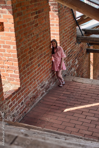 Young woman with brown hair in pink dress leans against red brick wall. She stands in sunlit alley under wooden beams, creating shadows and rustic atmosphere