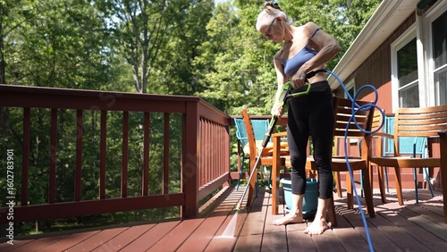 A mature woman is using a pressure washer to clean her wooden deck, ensuring it is free of dirt and grime while working in a bright garden setting.