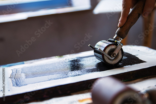 A closeup of an artist hand using a brayer to apply black ink onto a linocut surface in a linocut printmaking process.
