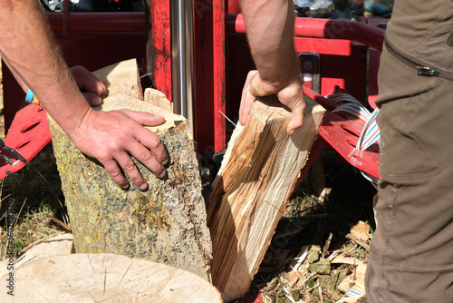 Log splitter in action splitting a thick log, with two people operating the machine outdoors on a sunny day.