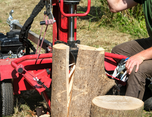 Log splitter in action splitting a thick log, with two people operating the machine outdoors on a sunny day.