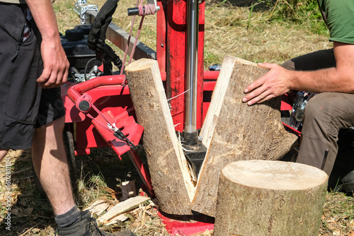 Log splitter in action splitting a thick log, with two people operating the machine outdoors on a sunny day.