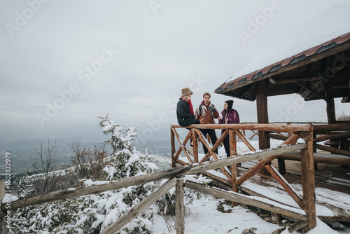 Фотография Friends convene atop a snowy mountain near a wooden shelter, sharing laughter and moments, surrounded by beautiful winter landscapes, enhancing the warmth and camaraderie of the scene