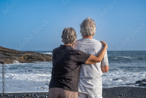 Rear view of elderly lovely couple hugging each other while standing by the seaside looking at the horizon over water. The ocean waves crash on the rocks in the background during golden hour