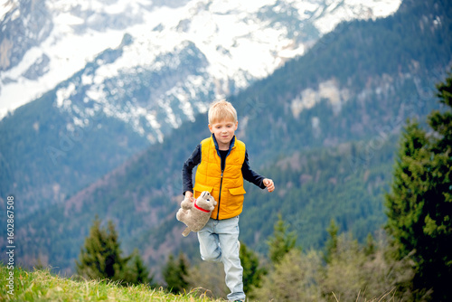 Little boy in bright yellow vest running on a green meadow in the Alps with snow-covered mountains in the background. Joyful childhood moment in the great outdoors. Family hiking vacation in the mount