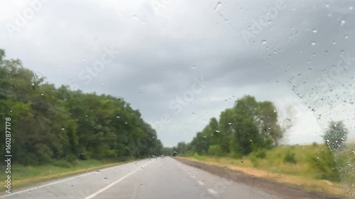 Car view on road with green trees and cloudy sky in rainy weather. Raindrop on glass.