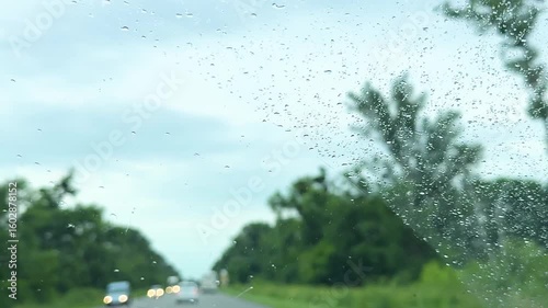 Raindrops on car window focusing side mirror, stormy atmosphere outside, concept of rainy travel, vehicle safety during bad weather. Side mirror reflecting road through rainy car window, overcast sky.
