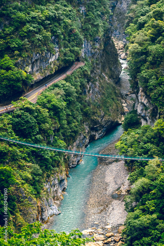 High-angle view of forest and river flowing through Taroko National Park, Taiwan. Clear blue water streams over rocks with moss in crevices of the mountain. There is a steel bridge across the trail.