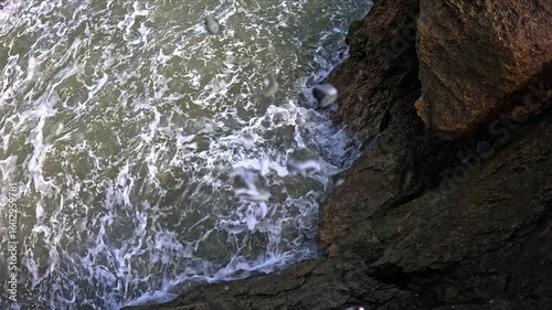 Bright sunlight reveals foaming sea crashing in slow motion, the camera moves into the water as waves hit the rocks.