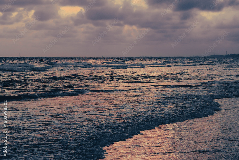 Obraz premium A serene sunset over a wavy ocean, reflecting purple and orange hues from the cloudy sky onto the water, with a distant coastline visible.