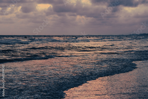 A serene sunset over a wavy ocean, reflecting purple and orange hues from the cloudy sky onto the water, with a distant coastline visible.