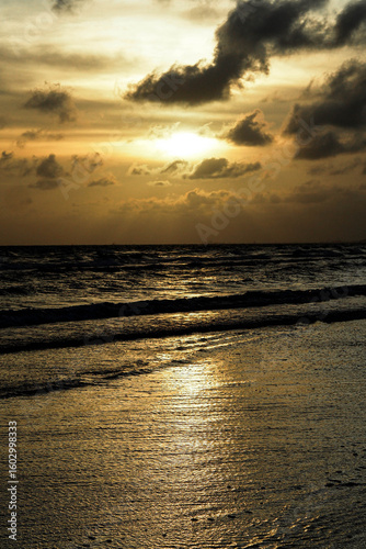 A golden sunset over a calm beach with dark waves reflecting the light and clouds overhead.