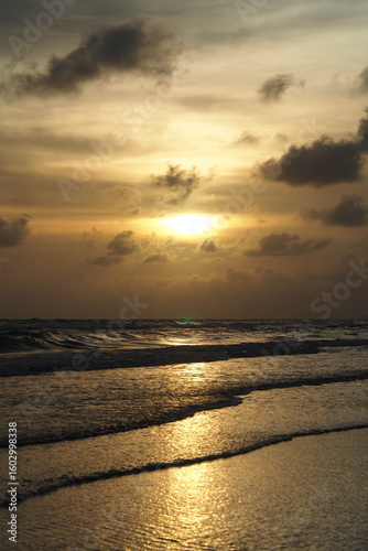 A dramatic sunset over the ocean with the sun's reflection on the waves and wet sand, under a cloudy sky.